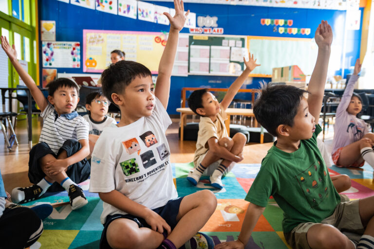 Students sitting on carpet raising their hands in class